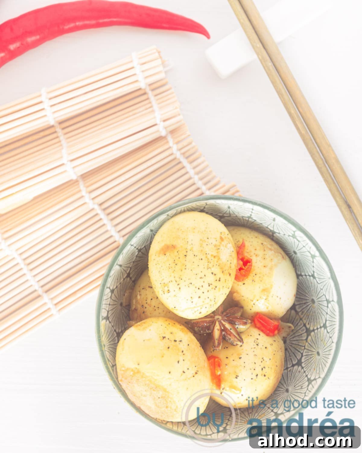A photo from above. A white background. Bottom right a green dish filled with stewed eggs, peppers and star anise. Linsboen a bamboo mat and a red pepper. Top right two golden chopsticks