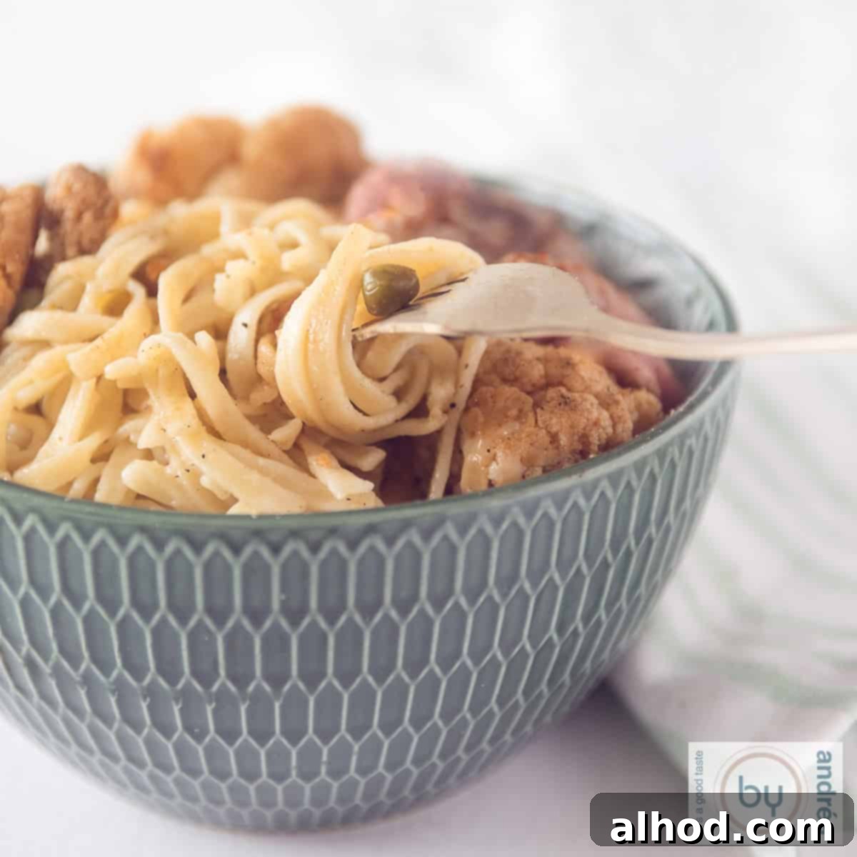 A square photo of a green bowl filled with cauliflower, peas, steak and noodles. A fork with some noodles twisted around it
