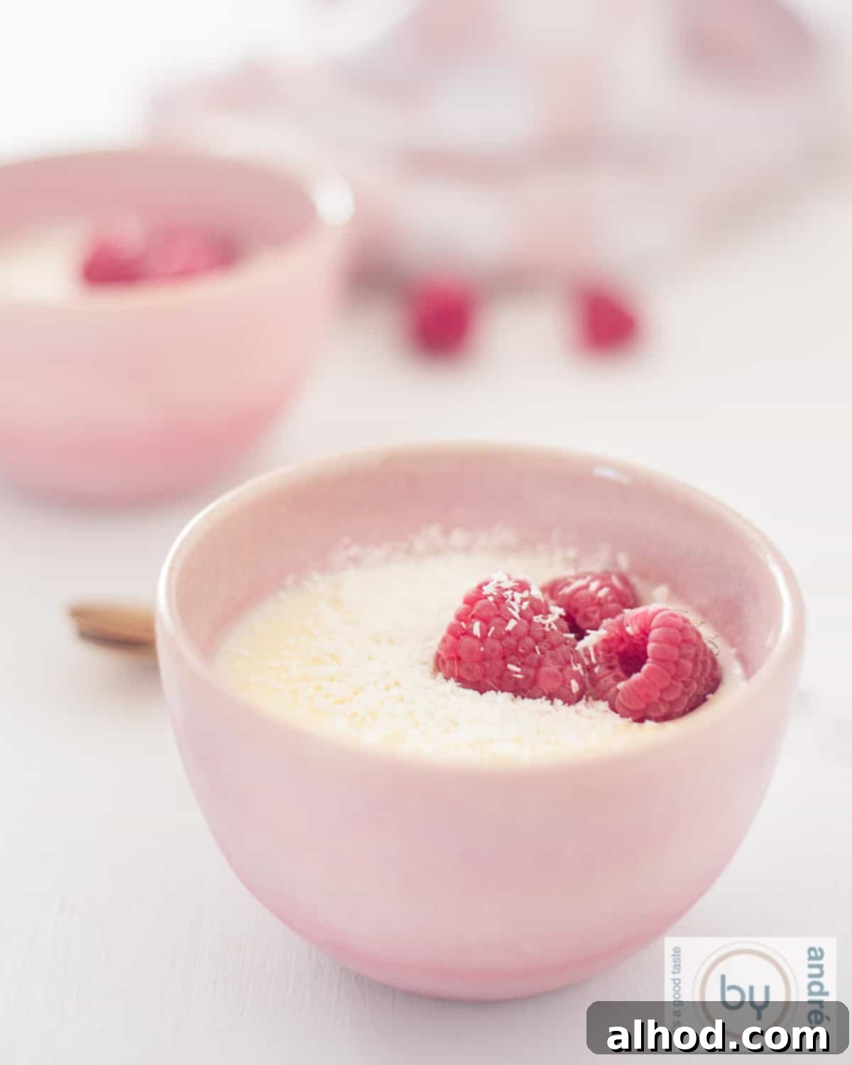 Easy 3-Ingredient Creamy Lemon Custard 2 A height photo of two pink bowls diagonally behind each other filled with lemon custard, coconut and raspberry on a white background