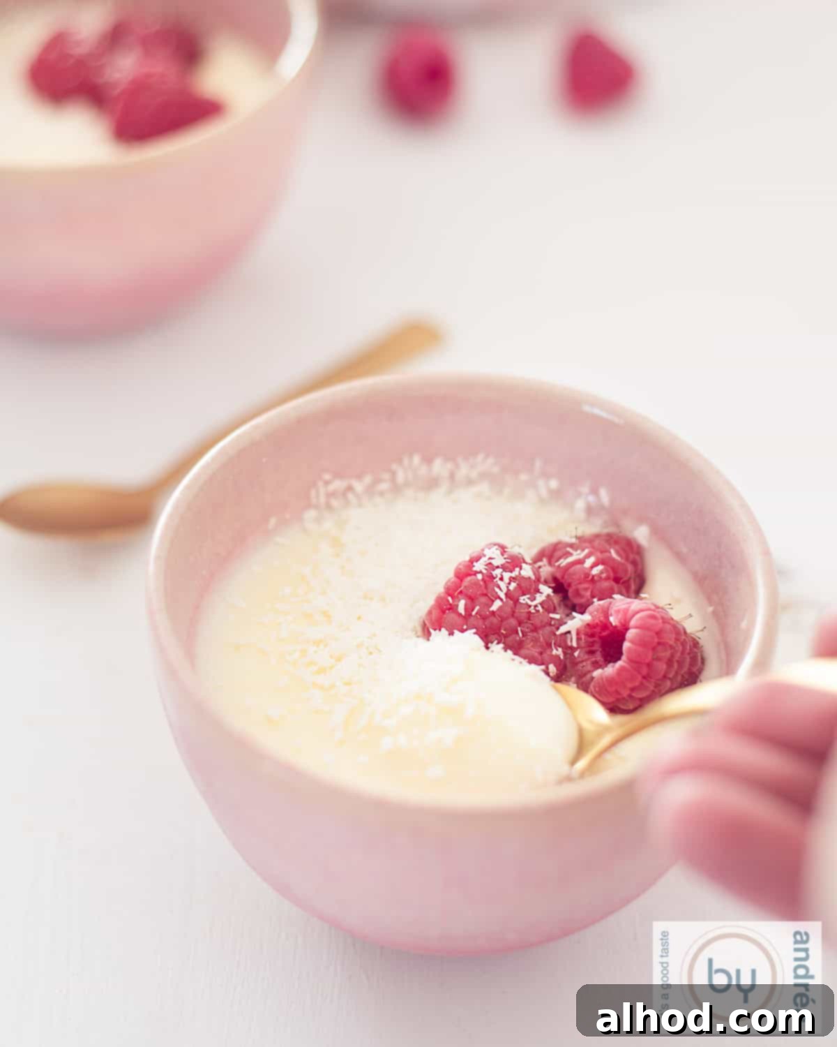 Easy 3-Ingredient Creamy Lemon Custard 8 A height photo with two pink bowls diagonally behind each other with lemon cream custard, raspberries and coconut. A spoon takes a scoop.