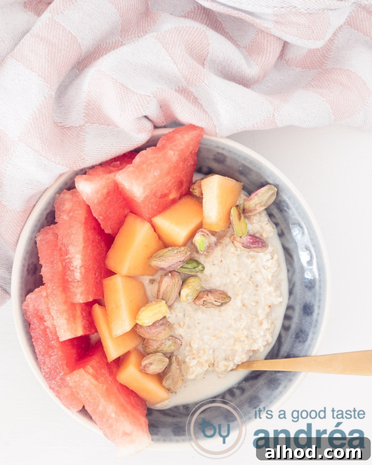 Melon Medley Overnight Oats 2 A photo from above with a gray bowl filled with overnight oats with two types of melon and pistachio on a white background. At the top a pink white cloth.