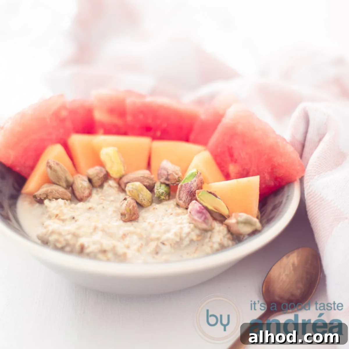 Melon Medley Overnight Oats 6 A square photo with a gray bowl with oatmeal, pistachio, two kinds of melon on a white background. A pink white tea towel in the background