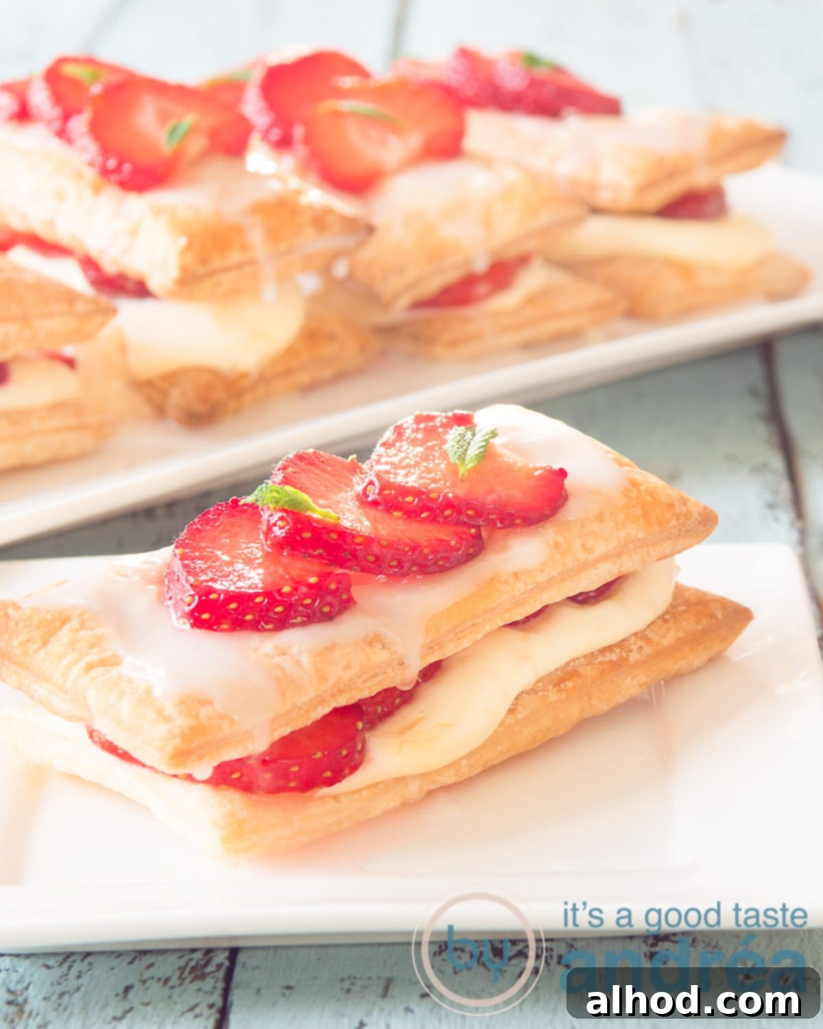 Decadent Strawberry Napoleon 5 A delicate puff pastry strawberry pastry on a white plate with a blurred blue background, showing more similar pastries.