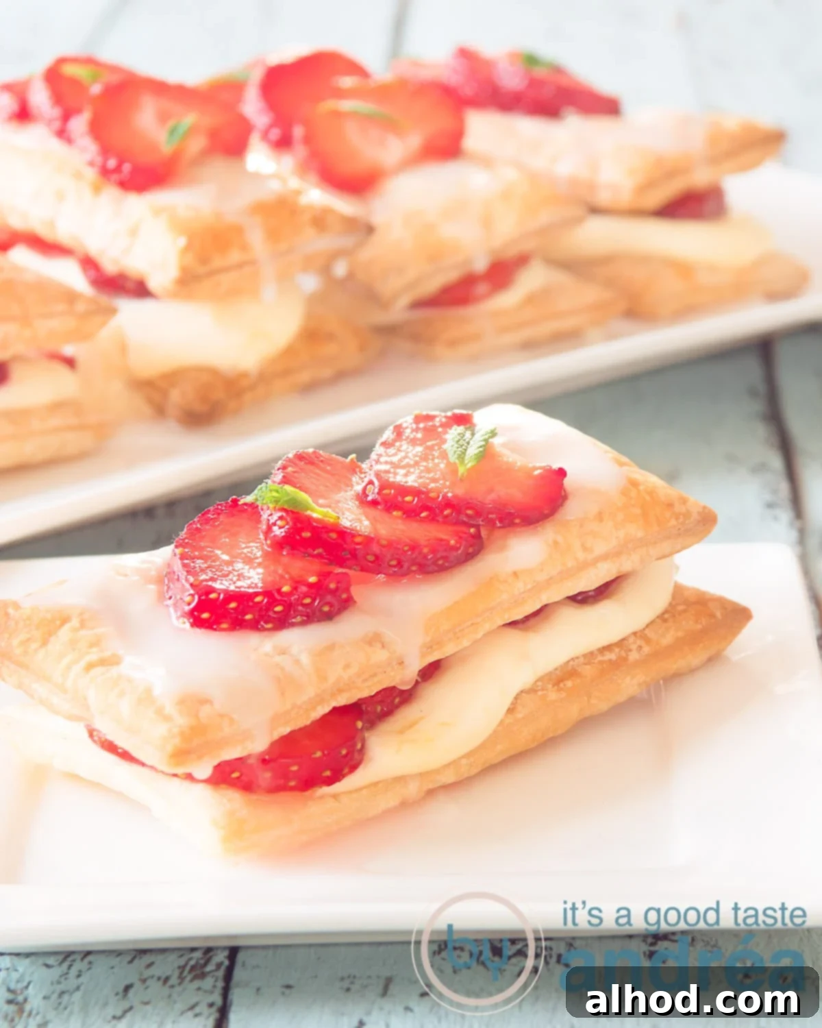Elegant Strawberry Napoleon Pastry 5 A delightful puff pastry strawberry dessert sits on a white plate, with a soft blue background. More pastries are visible in the blurred background, suggesting a delightful array of treats.