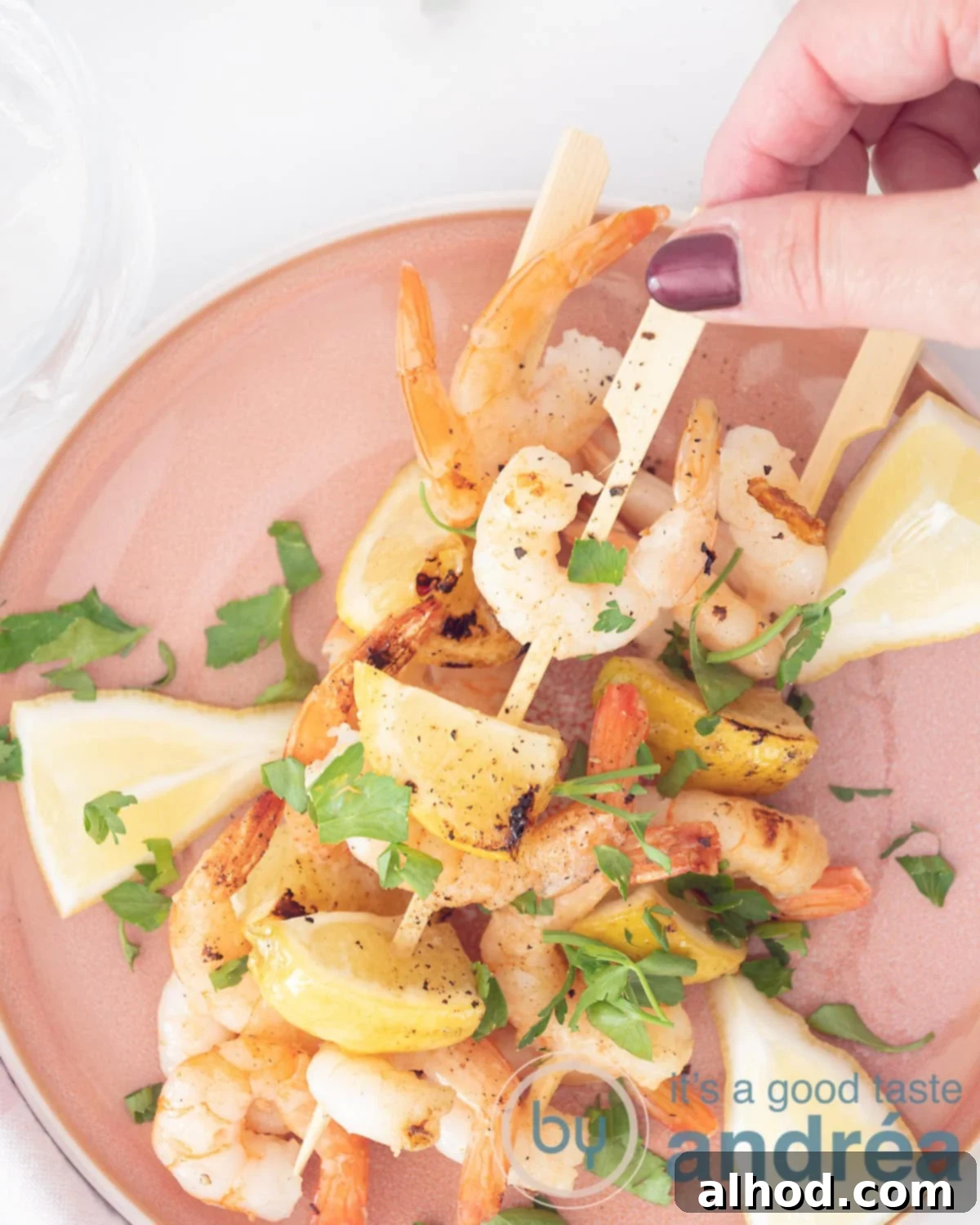 Grilled Lemon-Garlic Shrimp Skewers 2 A photo from above with three king prawn skewers. A hand grabs the middle skewer of the pink plate. Sprinkled with parsley. A white background.