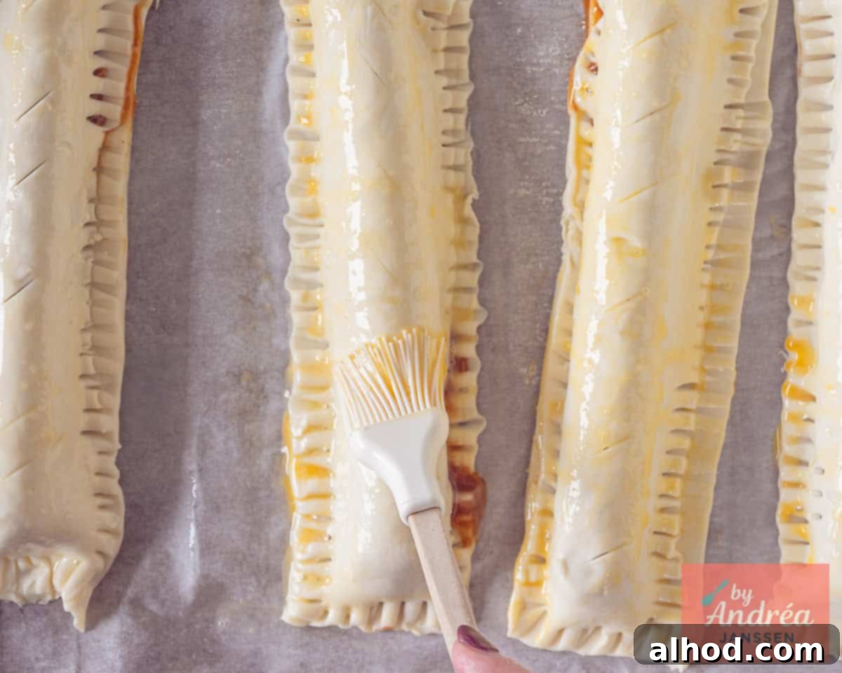 Freshly assembled frikandel rolls on a baking sheet, being brushed with a beaten egg for a golden finish.