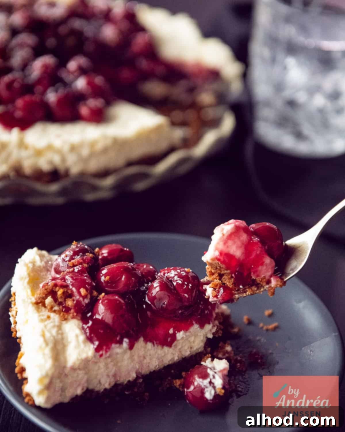 A slice of creamy Dutch Monchoutaart with a distinct Biscoff crust, fluffy white cream cheese filling, and a glossy red cherry topping. A bite has been taken, revealing the perfect layers.