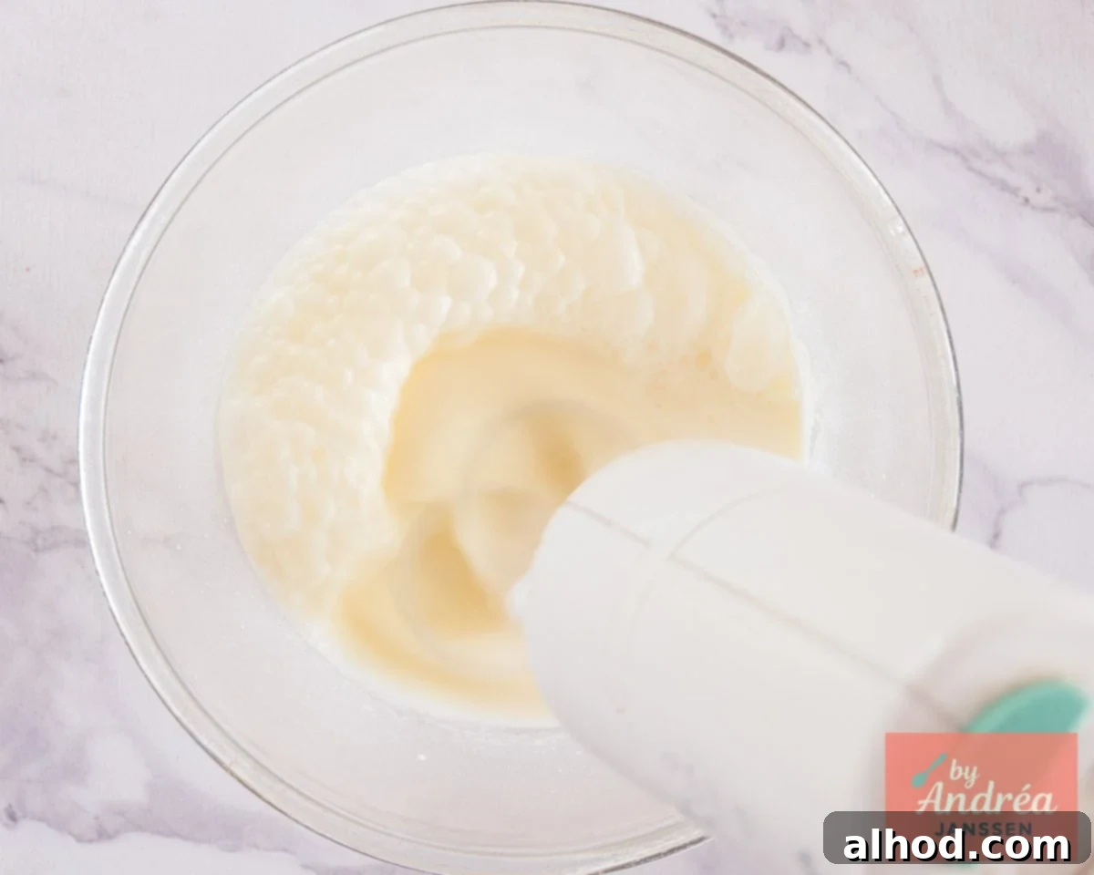A glass bowl with heavy cream being whipped to soft peaks using an electric mixer, creating a light and airy consistency.