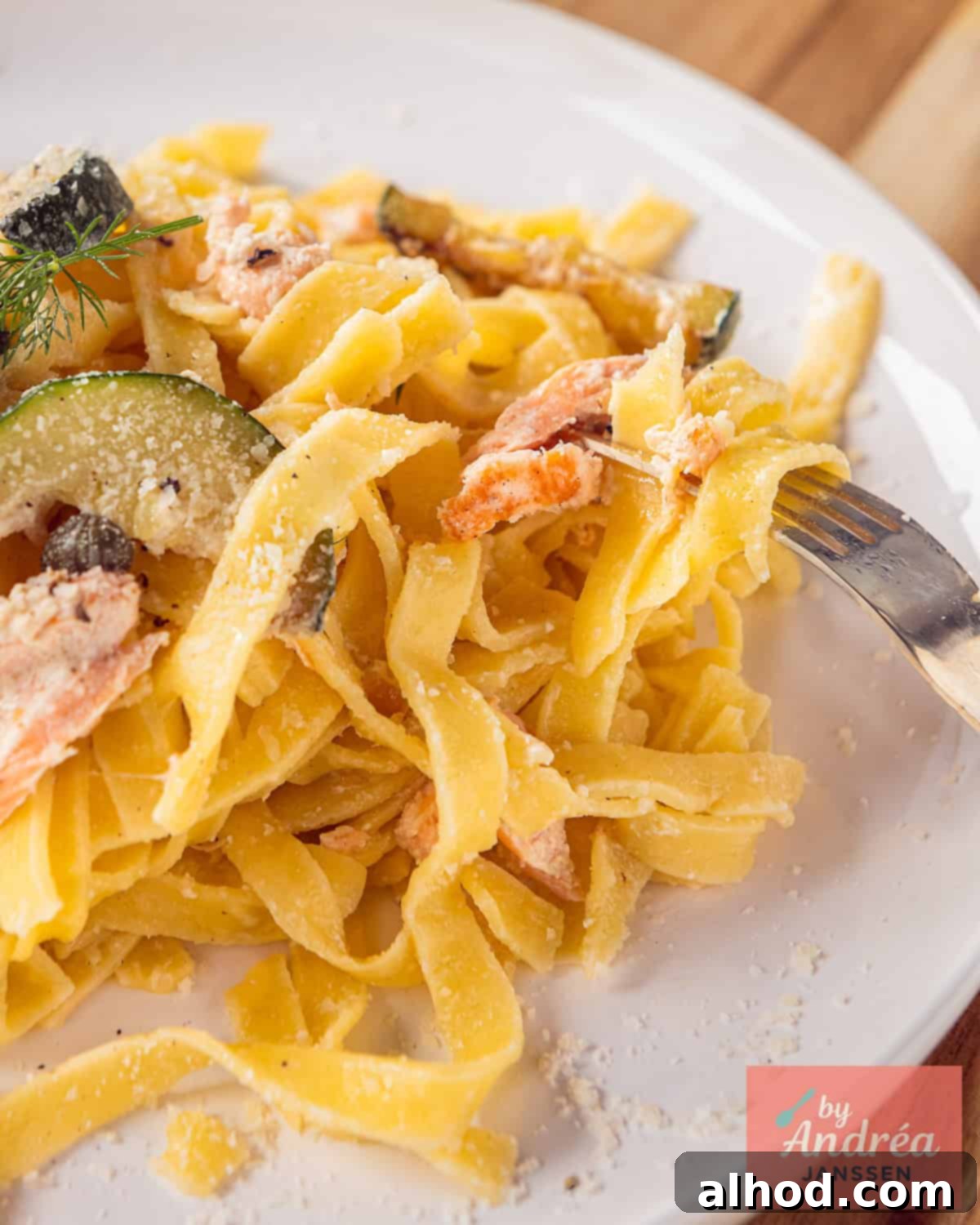 A portion of a white plate with pasta, salmon, zucchini, capers and parmesan cheese.