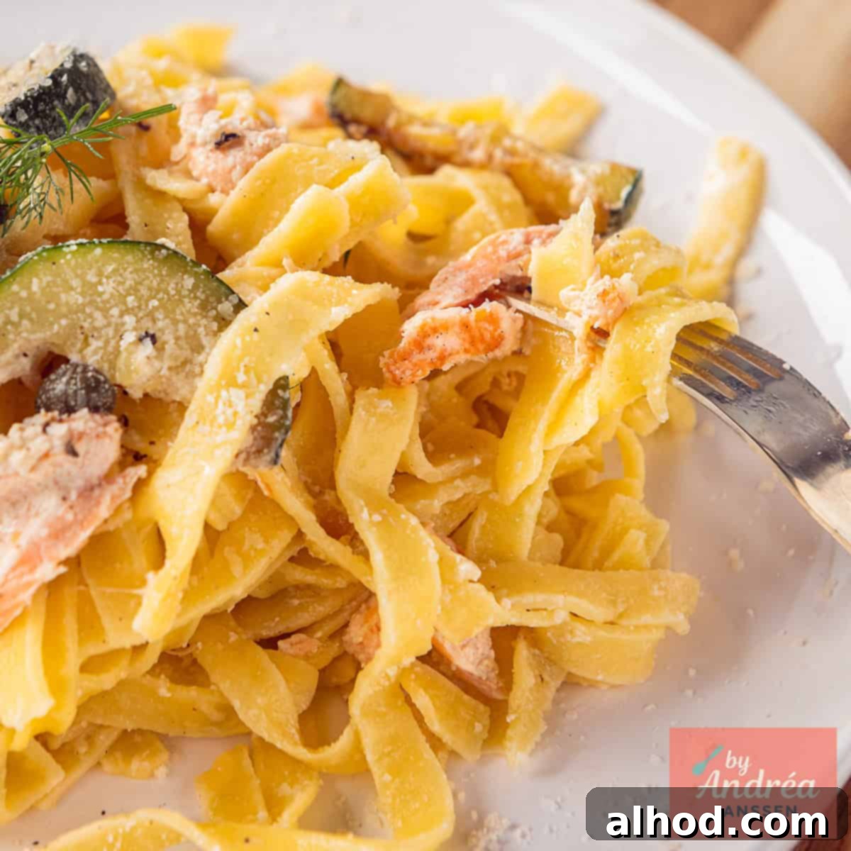 A square photo showing part of a white plate with pasta, zucchini, salmon, capers and creamy sauce.