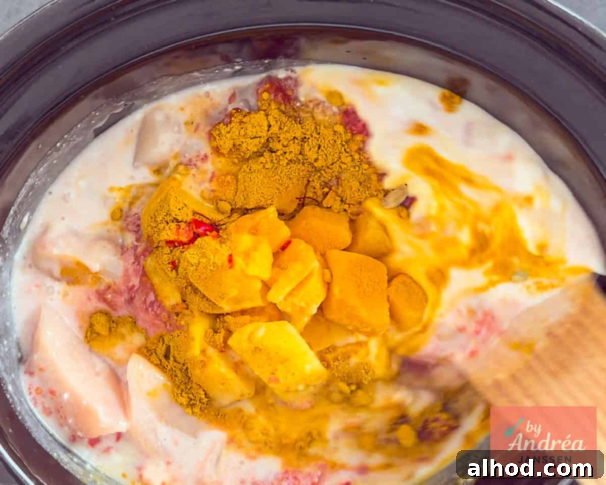 A slow cooker filled with chicken, tomatoes, coconut milk, and mango cubes, with a scattering of spices on top, ready for stirring.