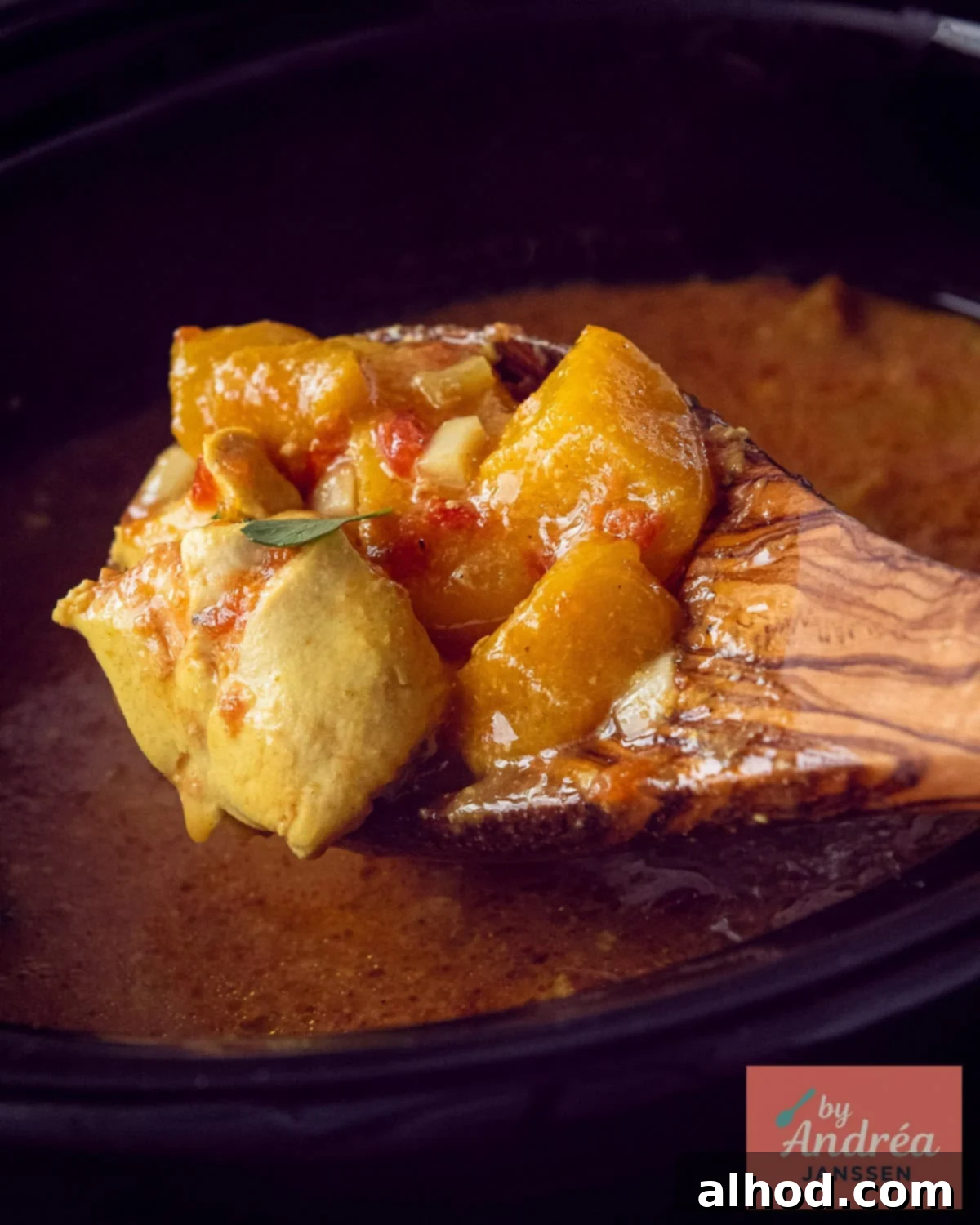 A wooden spoon scooping a portion of rich, creamy slow cooker chicken curry with tender chicken pieces and sweet mango from the pot.