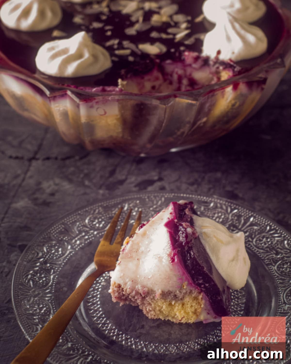 A piece of trifle with sherry, red fruit, and custard on a glass plate. The rest of the trifle is in the background.