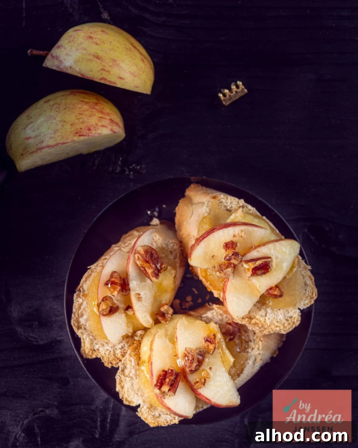 An overhead photo of three slices of baguette with apple, walnut, brie and honey on a dark background.