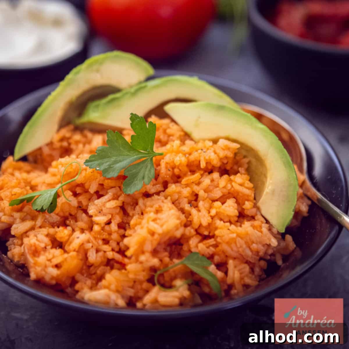 A square photo with a black bowl of red Mexican rice with avocado and parsley